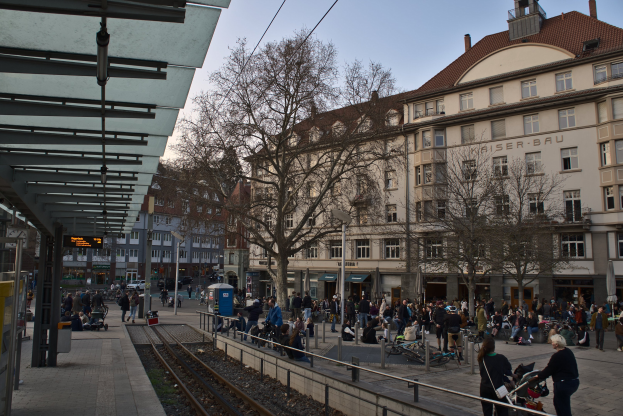 Eine belebte Straßenkreuzung mit einem Bahnhof auf der linken Seite, umgeben von Gebäuden, Bäumen, Laternen und Texttafeln, mit Menschen, die gehen, auf Bänken sitzen und stehen.