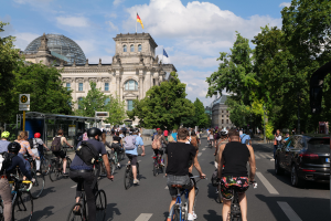 Eine Gruppe von Menschen, die Fahrräder auf einer von Bäumen gesäumten Straße in Berlin fahren, mit Gebäuden auf beiden Seiten, einer Bushaltestelle auf der rechten Seite, einem bewölkten Himmel und einer Flagge, die auf einem der Gebäude weht.