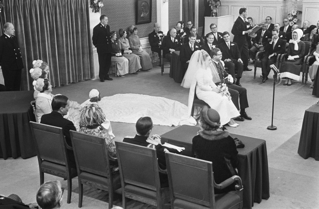 Schwarzes und weißes Foto einer Hochzeitszeremonie mit sitzenden und stehenden Menschen, einem Mann mit Mikrofon, einer Frau mit einem Bukett, einem Podium mit Mikrofon, Blumenschmuck, einem Vorhang, einem gerahmten Foto an der Wand und einer Tür im Hintergrund.