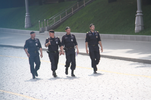 Drei Polizisten in Uniform und mit Mützen gehen eine Straße mit Pfählen, Geländern, Treppen und Gras im Hintergrund entlang.