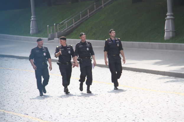 Drei Polizisten in Uniform und mit Mützen gehen eine Straße mit Pfählen, Geländern, Treppen und Gras im Hintergrund entlang.