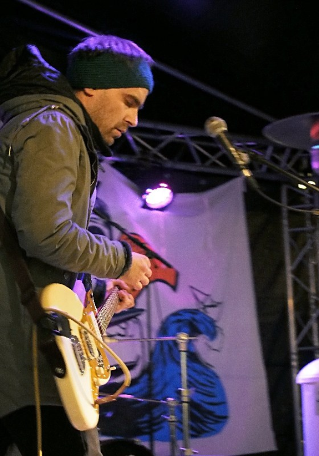 Ein Mann in einer Jacke und einer Mütze spielt eine E-Gitarre auf der Bühne mit einem Mikrofon vor sich, ein Schlagzeug zu seiner Rechten, Bühnenlichter und ein Banner im Hintergrund.