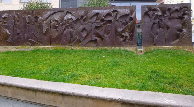 Große Metallskulptur der Berliner Mauer auf einer grasbewachsenen Fläche umgeben von Bäumen und Gebäuden mit Fenstern im Hintergrund.